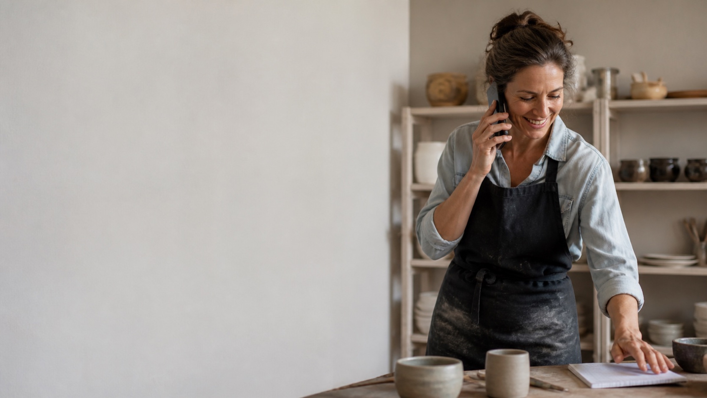 A small business owner taking an inquiry call in her workshop, smiling as she reaches for a notepad